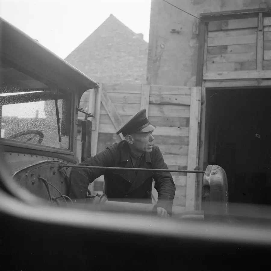 Black-and-white photograph of a driver in a peaked cap leaning out beside his vehicle