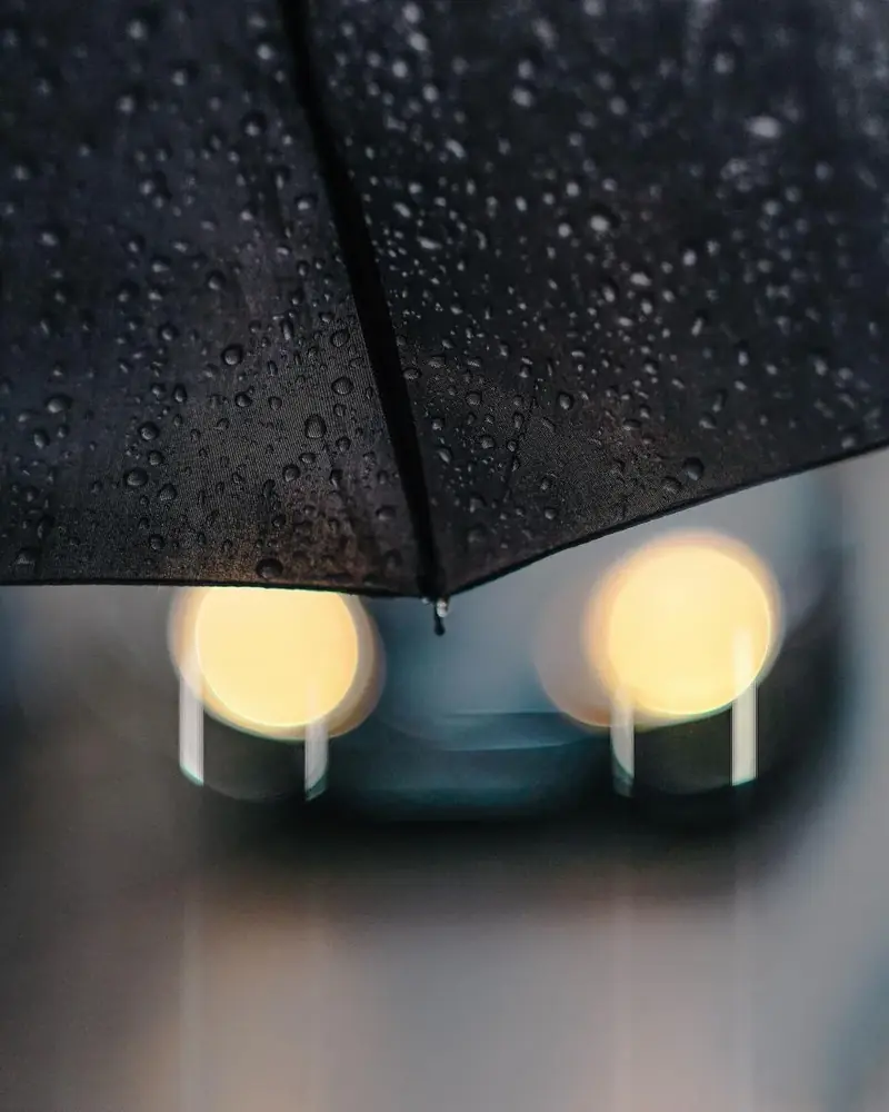 A black umbrella held over the glowing headlights of a waiting car on a wet evening